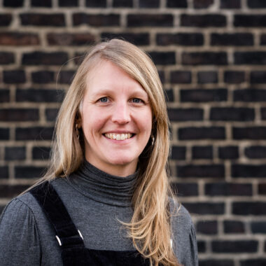 Joanna Mills stands in front of a brick wall at Spode factory, wearing a grey turtleneck and dungaree dress.