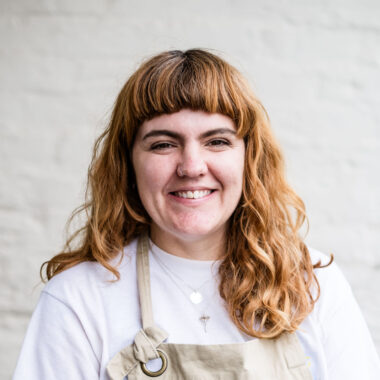 Alice Thatcher is a white woman with reddish brown hair. She has thick wavy hair and a fringe and is wearing a white top and a tan apron with a BCB logo on it.
