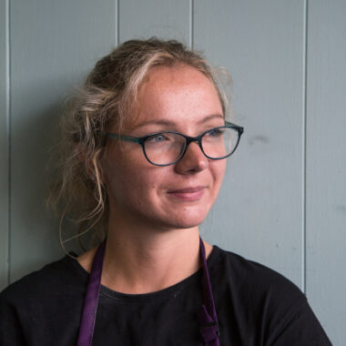 Joanna Dawidowska is standing in front of a green, panelled wall wearing a black top and glasses.