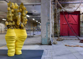 A tall ceramic sculpture with a yellow glaze standing in front of ceramic chains which are hanging from the ceiling.