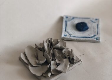 Two clay tiles are displayed on cream paper. One has handmade rose petals attached and the other is painted white with a blue shell attached in the centre.