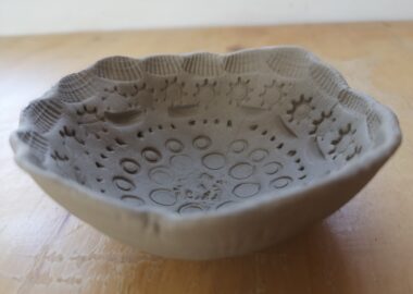 A press moulded clay bowl sits on a wooden table with patterns imprinted on the surface.