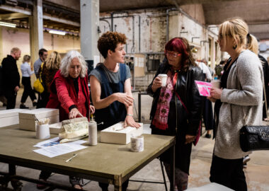 Artist Sarah Fraser is having a conversation with a visitor to British Ceramics Biennial 2019. She is causally leaning on a white ceramic mould.