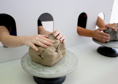 Two people rest their hands on two lumps of clay, unable to see what's in front of them.