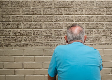 An artist sits in front a large clay wall with his back to the camera, writing poetry into the clay.