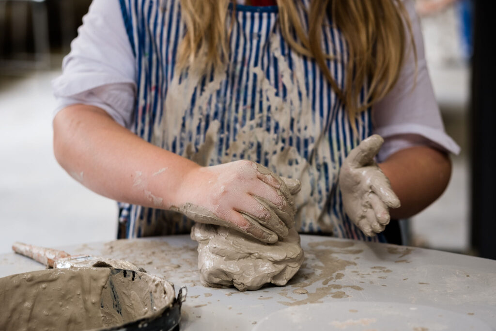 A school child sculpts with wet clay on a grey table.