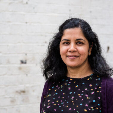 Associate Artist Zeba Imam is smiling at the camera, standing in front of a white brick wall.