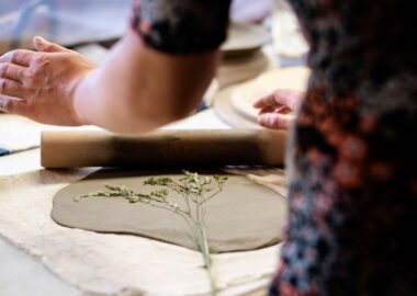 A person presses dried flower stems into a slab of grey clay to leave a mark.