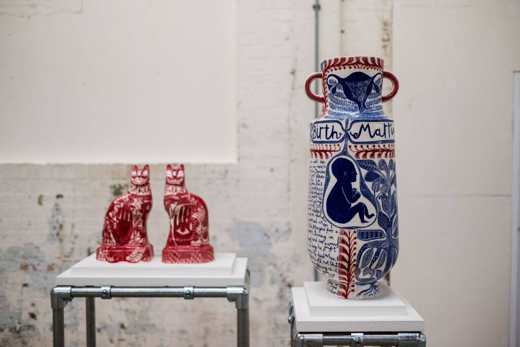 Two ceramic cats and a tall vase are displayed on plinths against a white background. The ceramics are illustrated with drawings.