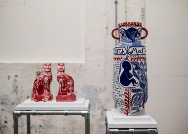 Two ceramic cats and a tall vase are displayed on plinths against a white background. The ceramics are illustrated with drawings.