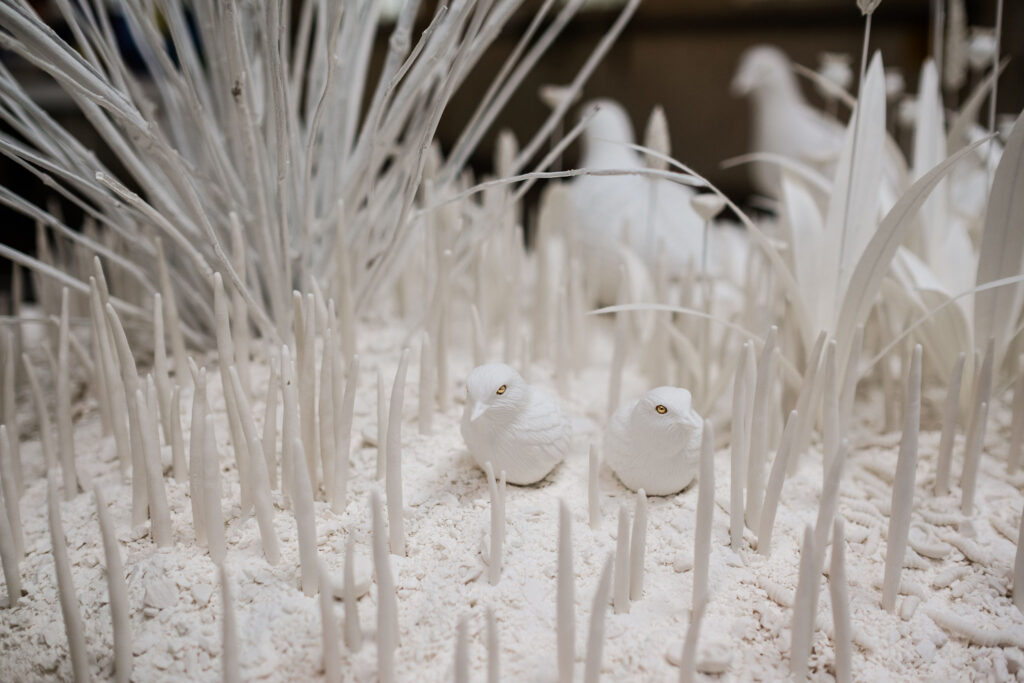 Two porcelain white birds sit atop white porcelain soil surrounded by white porcelain grass stems.