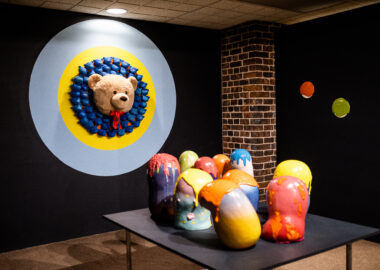 A collection of colourful ceramics sculptures are displayed on a black plinth infront a a teddy bear's, wall-mounted head.