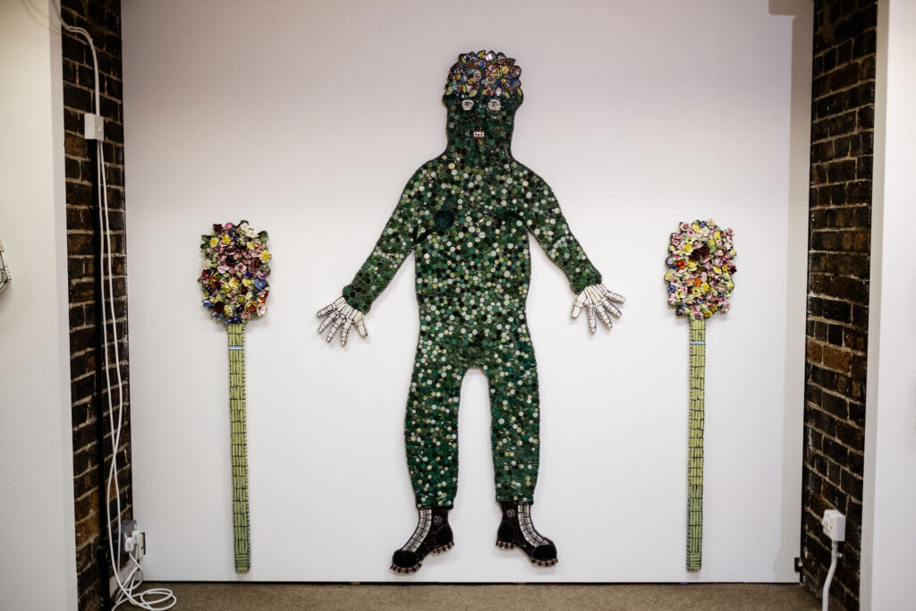 Mosaic figure of a man covered in leaves, with mosaic plants to either side of him, by Cleo Mussi mounted to a wall, AWARD Exhibition at British Ceramics Biennial 2021