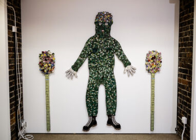 Mosaic figure of a man covered in leaves, with mosaic plants to either side of him, by Cleo Mussi mounted to a wall, AWARD Exhibition at British Ceramics Biennial 2021