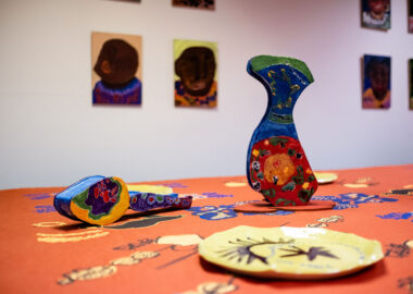 Colourful ceramic vases and a plate are displayed on a hand printed textile cloth with a backdrop of painted portraits by artist Mawuena Kattah
