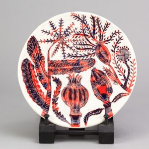 A white ceramic plate with red illustrated seed heads made by Vicky Lindo and William Brookes is displayed on a dark brown plinth.