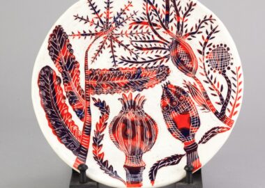 A white ceramic plate with red illustrated seed heads made by Vicky Lindo and William Brookes is displayed on a dark brown plinth.
