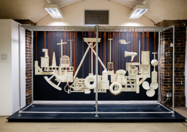 A selection of ceramic objects suspended in the air by orange string. Each object, rocking horse, life jacket, a noose, reflects a part of the experience of emigration