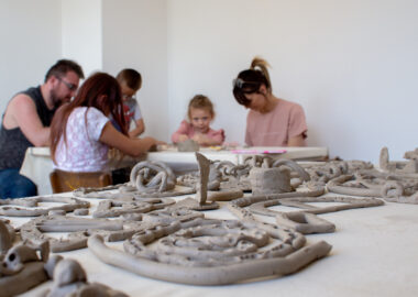 A number of coiled ceramic shapes are laid out on a table with a group of people making on a table in the background.