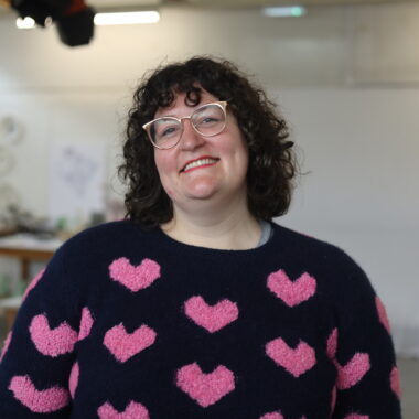 Alex is smiling and standing in a white room, in front of a ceramics exhibition.