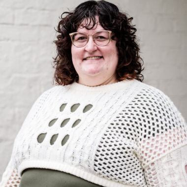 Alex Rabishaw is a woman with pale skin and brown curly hair. She is wearing a green dress and white crochet sweater.