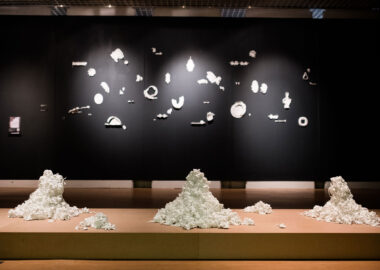 An art exhibition space. A platform on the floor has 3 piles of ceramic flowers, and the wall behind features white ceramic pieces on a black wall.