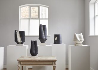 A bright with gallery with five vessels on wooden pedestals. The vessels are cylindrical, tapering inwards towards the top and have long creases going up the form.