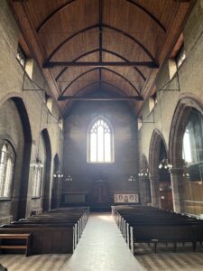 View from inside All Saints Church, looking across the rows of pews to an arched window on the back wall, and high vaulted ceilings with rafters going across.