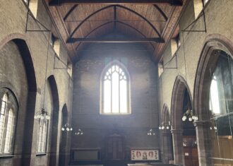 View from inside All Saints Church, looking across the rows of pews to an arched window on the back wall, and high vaulted ceilings with rafters going across.