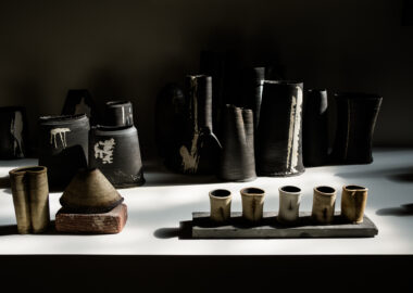 Cluster of black clay vessels behind a line up of small clay cups as part of Dan Kelly's work in the 2023 British Ceramics Biennial