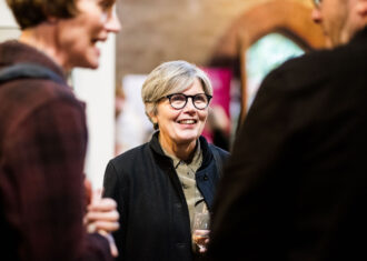 Rosy Greenlees, Chair of British Ceramics Biennial, stands talking with two people who are blurred in the foreground.