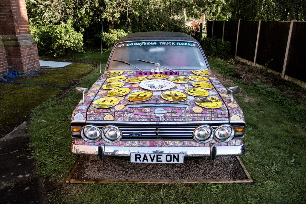 A car has been completely covered in a mosaic of pink tiles with yellow smiley face accents.