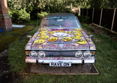 A car has been completely covered in a mosaic of pink tiles with yellow smiley face accents.