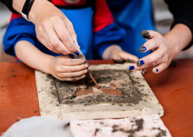 Someone is helping a child squeeze clay slip onto a clay tile in the Tactile Project Space.