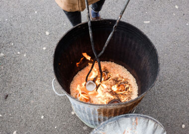 Small ceramic pieces sit in a metal barrel on top of some sawdust. The sawdust has begun to catch fire due to the temperature of the ceramic pieces. This is part of the firing process known as raku.