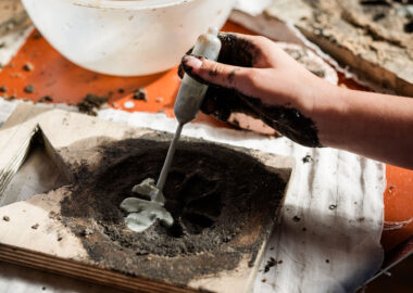 Close-up of a hand holding a pipette of clay slip and squeezing it into an indented pattern on a clay tile.