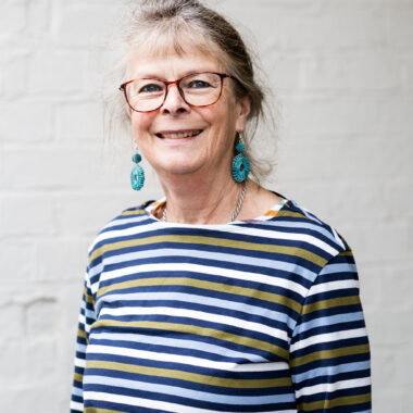 Sue Blatherwick has light skin and medium brown hair pulled into a ponytail. She is wearing bright turquoise earrings and a blue-striped shirt.