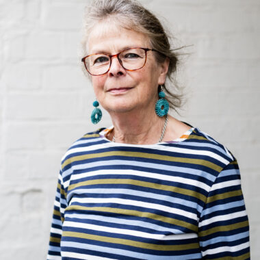 Sue Blatherwick has light skin and medium brown hair pulled into a ponytail. She is wearing bright turquoise earrings and a blue-striped shirt.