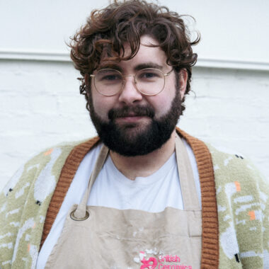 Matt Whiting has fair skin, short, curly dark brown hair and a full but close-cropped dark brown beard. They are wearing round gold wire-framed glasses and a cardigan with geese on it.