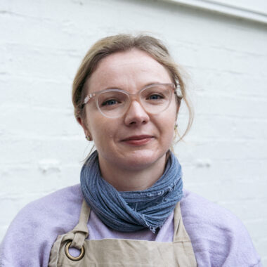Joanna Dawidowska has fair skin with dark blonde hair. She is wearing glasses, a blue scarf and a lilac top.