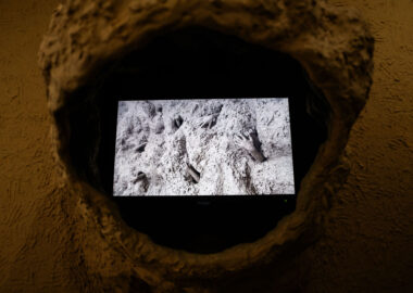 A screen within a clay hood showing hands poking out of a mound of wet clay.